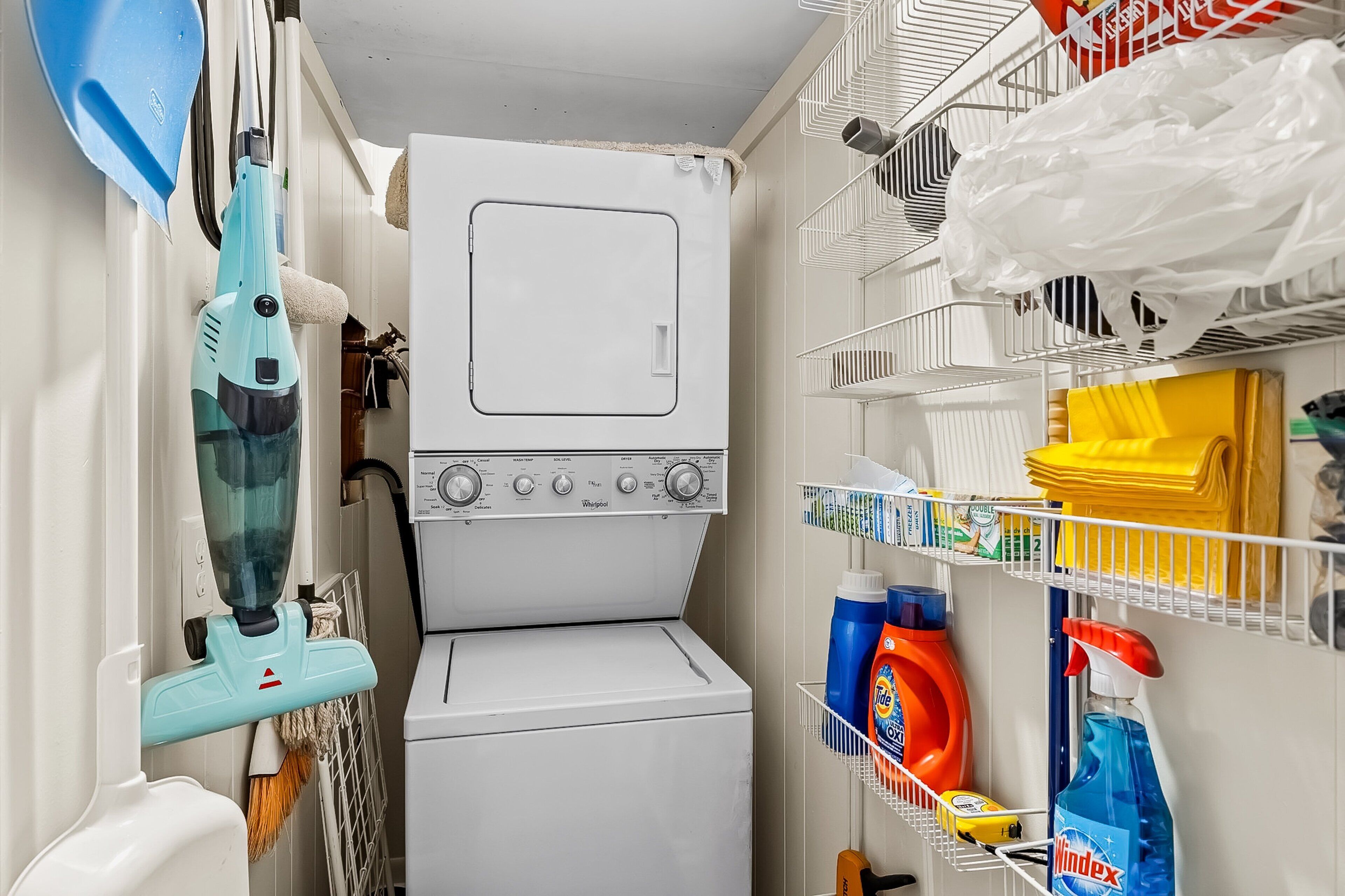 A compact laundry room with a front-loading washing machine, a blue handheld steamer, and shelves stocked with cleaning supplies.
