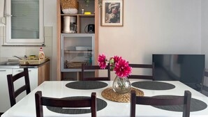 Un comedor con una mesa blanca, sillas de madera oscura, un jarrón con flores rosadas y un televisor a la derecha.