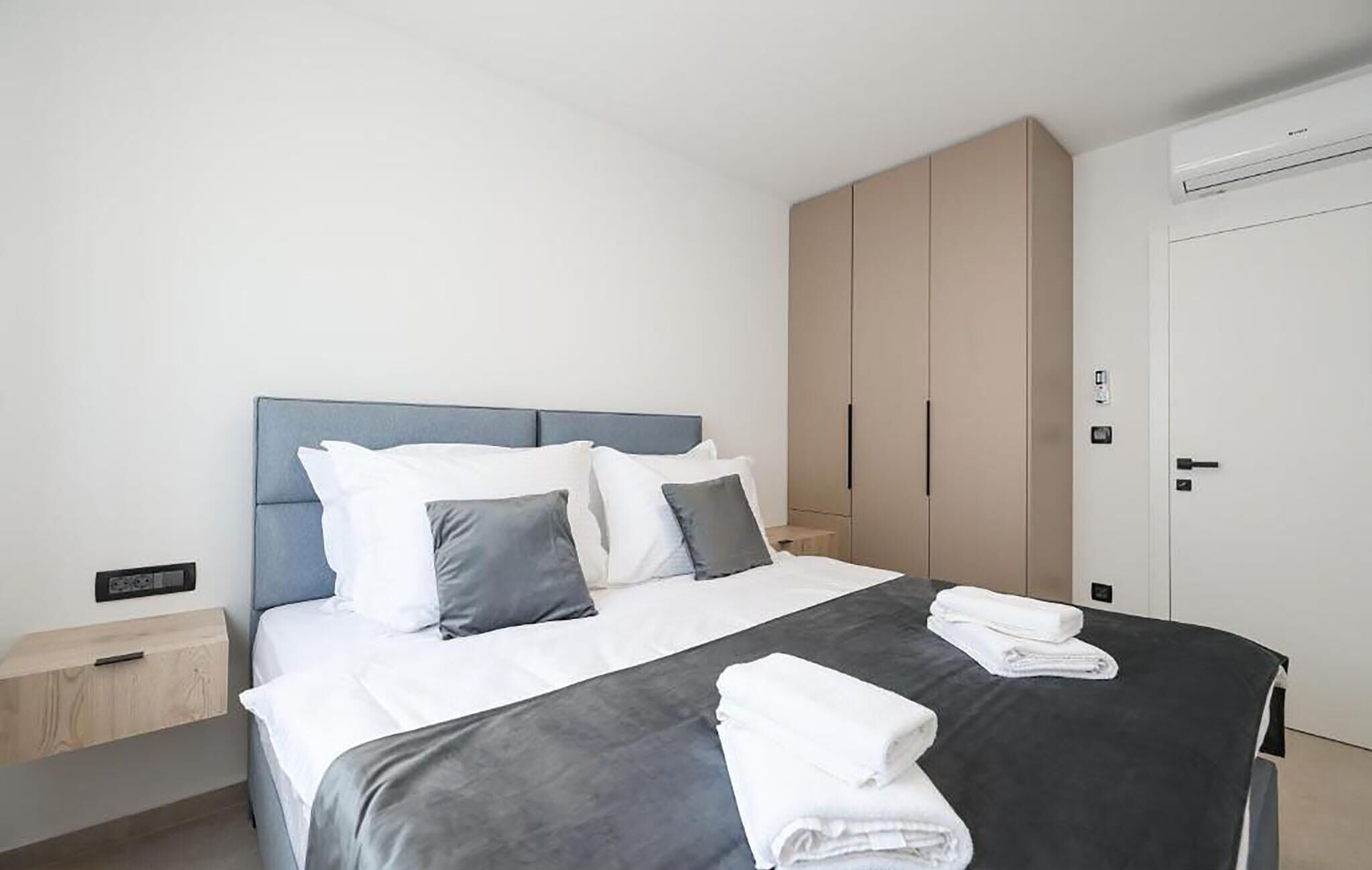 Una cama bien hecha con ropa de cama blanca y gris, una colcha gris oscuro y toallas blancas dobladas sobre la cama.