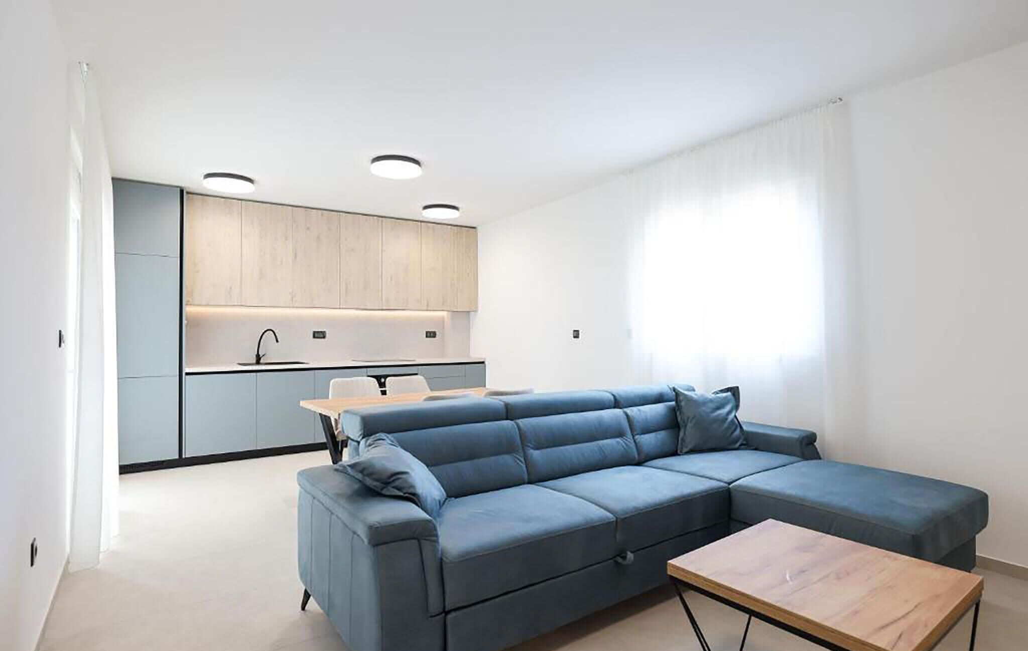 Ein modernes Wohnzimmer mit einem Ecksofa, einem kleinen Holztisch und einem Küchenbereich im Hintergrund.