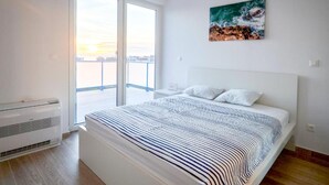 A modern bedroom with a large bed, a view of the city through a sliding door, and a painting of a wave on the wall.