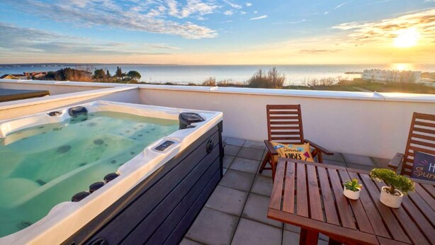 A rooftop terrace with a hot tub, wooden chairs, and a table with a potted plant.