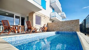 A pool area with a tiled edge, wooden lounge chairs, and a stone wall.