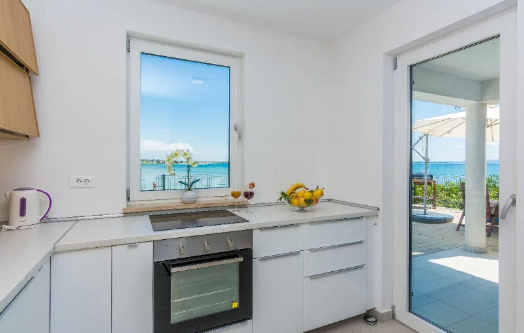 Une cuisine moderne avec vue sur la mer, une petite fenêtre et un saladier de fruits posé sur le plan de travail.