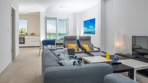 A modern living room with a grey sofa, two armchairs, a small table, and a TV. There is a balcony with a view of the sea.