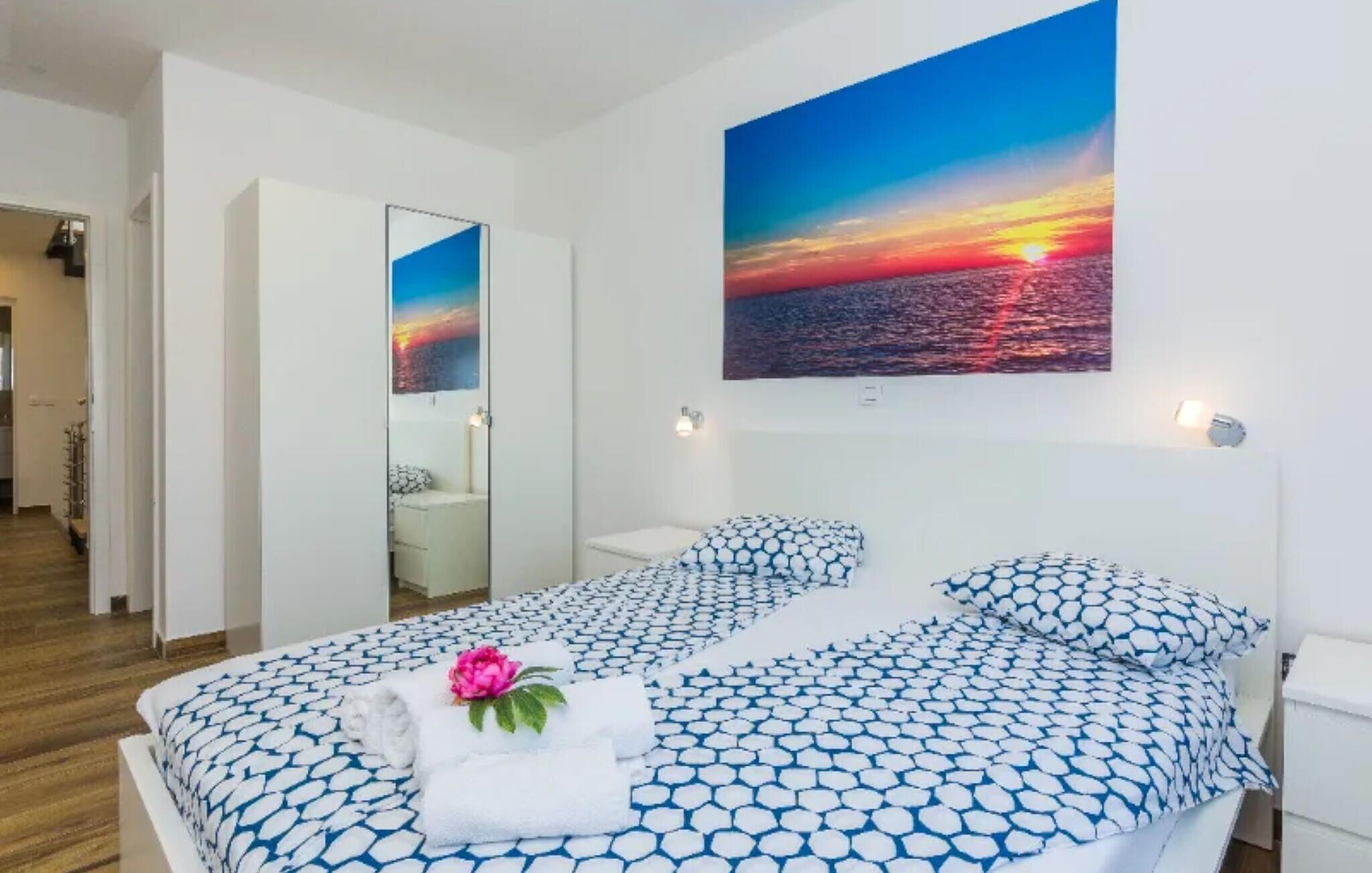 Une chambre à coucher avec un lit, un miroir et un tableau représentant un coucher de soleil sur l’océan.