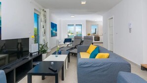 A modern living room with a flat-screen TV, a sofa with colorful cushions, a coffee table, and a dining area with chairs. - Lovely home in Privlaka (Privlaka)