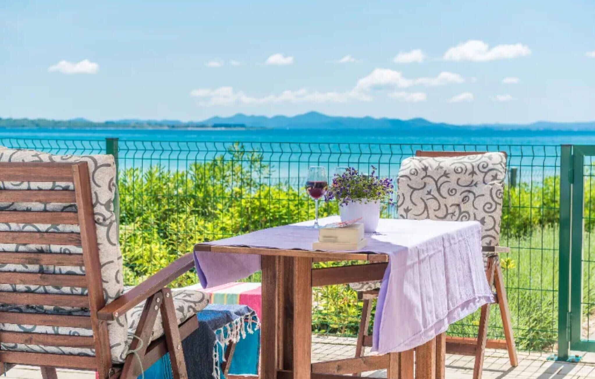 A table set with a wine glass, a book, and a potted plant, overlooking a scenic view of the sea and mountains.
