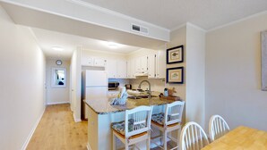 A kitchen with white cabinets, a granite countertop, and a dining area with a wooden table and white chairs. - 2nd-Floor Condo in Oceanfront Location w/WiFi, Screened-In Balcony, Shared Pool (Atlantic Beach)