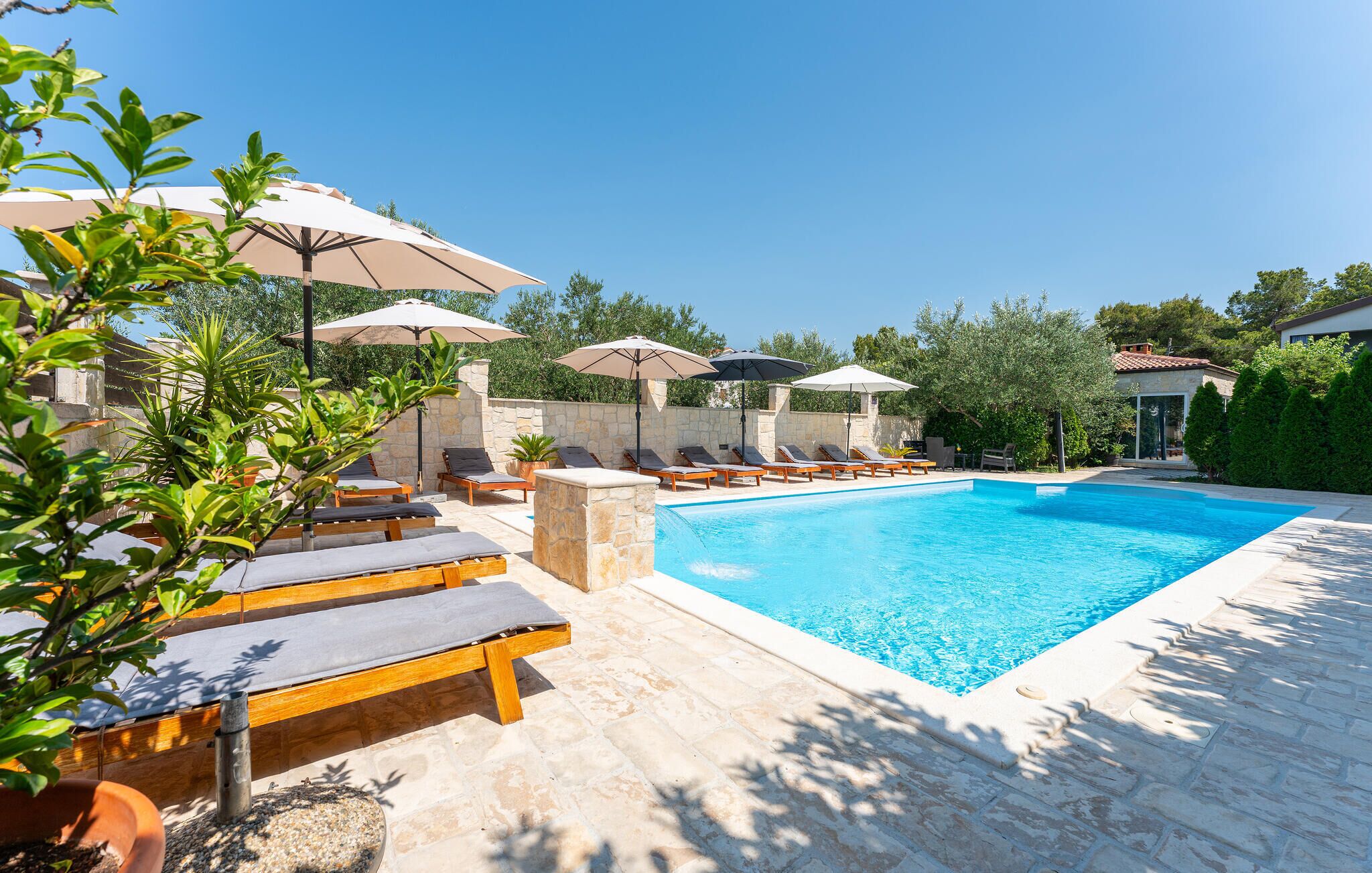 A poolside area with lounge chairs and umbrellas, a stone wall, and a clear blue pool.