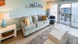 Un salon avec un mur bleu clair, un canapé gris agrémenté de coussins, une petite table d’appoint blanche avec une lampe, un pouf beige et une vue sur un balcon aménagé avec du mobilier d’extérieur.