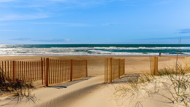 Una playa arenosa con una valla de madera que conduce al océano, y una persona caminando a lo lejos.