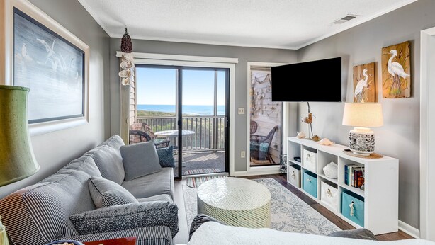 A modern living room with a sofa, coffee table, and a TV on a stand. There is a balcony with a view of the ocean. - 3rd Floor Oceanfront Condo w/Great Ocean and Pool Views and Shared Pools (Atlantic Beach)