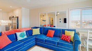 A modern living room with a blue sectional sofa, a white desk, and a large window with blinds. - Condo with on-site beach access, pools, grill/picnic areas, & private balcony (Atlantic Beach)