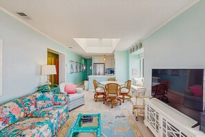 Ein Wohnzimmer mit Sofa, Couchtisch, Esstisch mit Stühlen, Fernseher und einer Küche im Hintergrund.