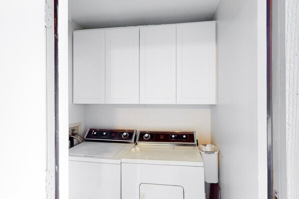A compact laundry room with a washing machine and dryer, white cabinets above, and a small trash bin to the side.