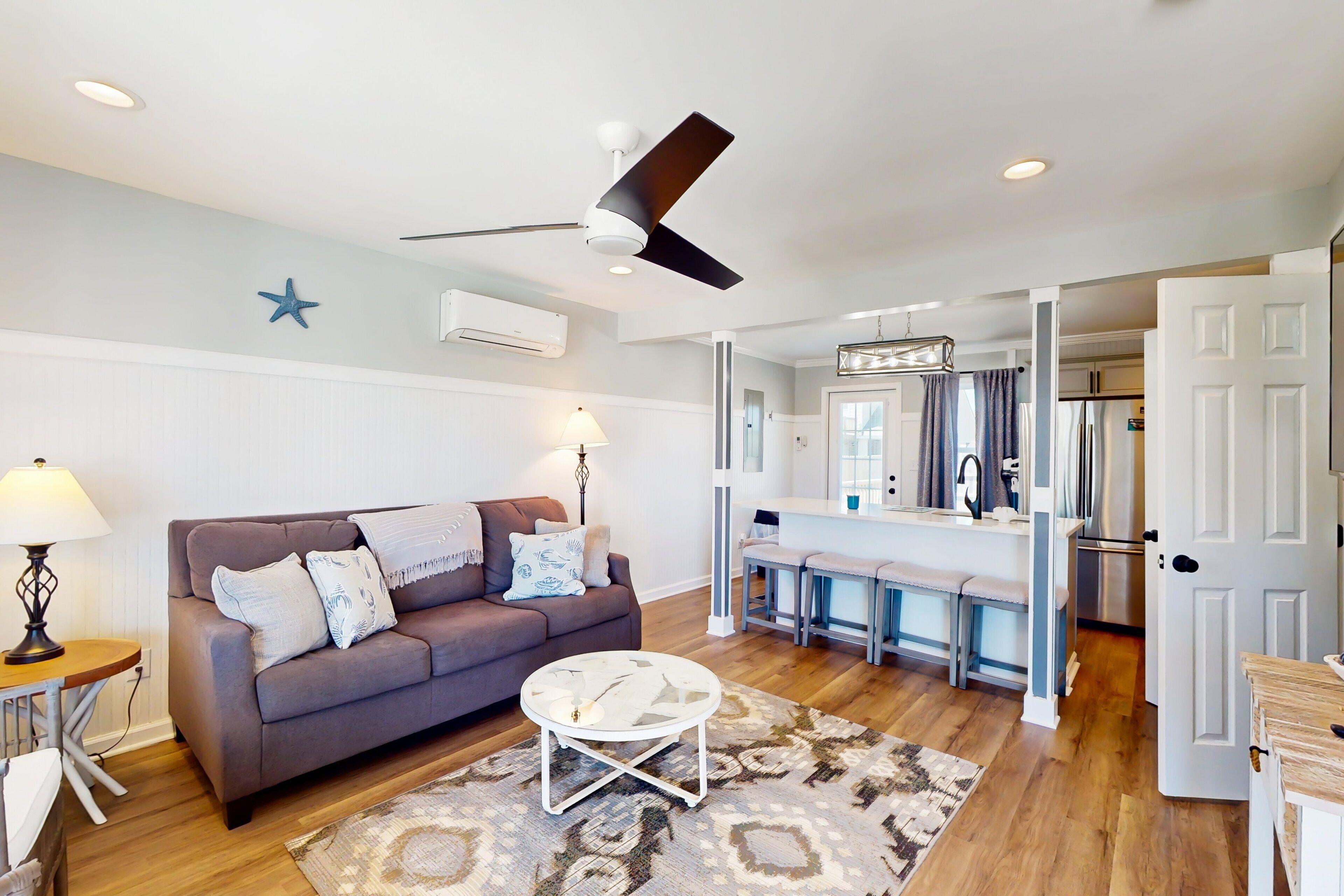 A modern living room with a sofa, coffee table, and a ceiling fan. A kitchen area is visible in the background with a bar counter and appliances.