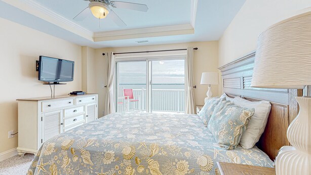A bedroom with a bed, a TV, a ceiling fan, a balcony, and a view of the ocean.