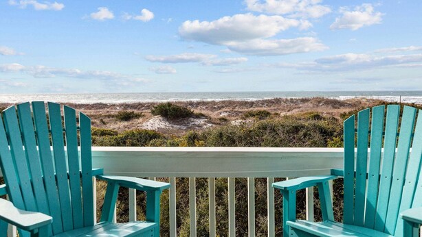 Twee turquoise Adirondack stoelen op een wit terras met uitzicht op een strand met duinen en een heldere lucht.