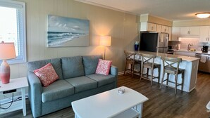 Un salon avec un canapé gris, une table basse blanche et un tableau représentant l’océan. On y trouve également un coin cuisine avec des meubles en bois et un tabouret de bar.