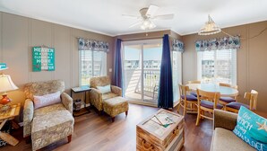 A spacious living room with a balcony, a ceiling fan, and a view of the water. - 3rd-Floor Condo Overlooking Courtyard w/Beach Views, WiFi, Shared Pools/Hot Tub (Atlantic Beach)