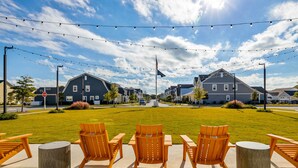 An outdoor seating area with wooden chairs and tables, a grassy lawn, and a row of houses in the background. - The Gough Coast; 3BR with seasonal pool access & community dock (Beaufort)