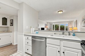 A modern kitchen with white cabinetry, a stainless steel dishwasher, and a large window providing natural light. - Beautiful oceanfront condo in the heart of Pine Knoll Shores with pool (Pine Knoll Shores)