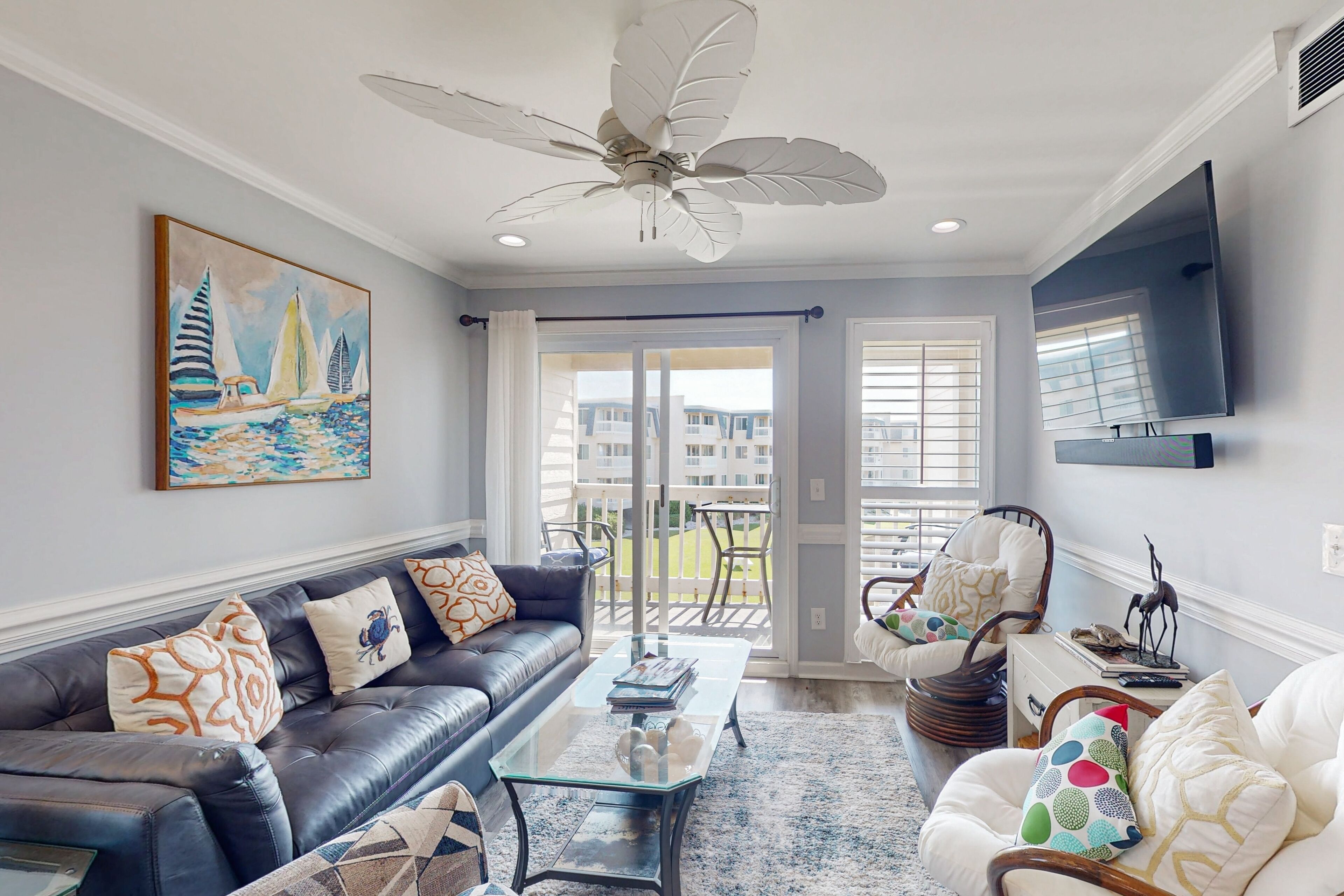 A living room with a black leather sofa, a glass coffee table, and a white armchair. There is a ceiling fan, a painting of sailboats, and a large window with blinds.