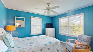 Un dormitorio con cama, una silla, cómoda, televisor y ventilador de techo.