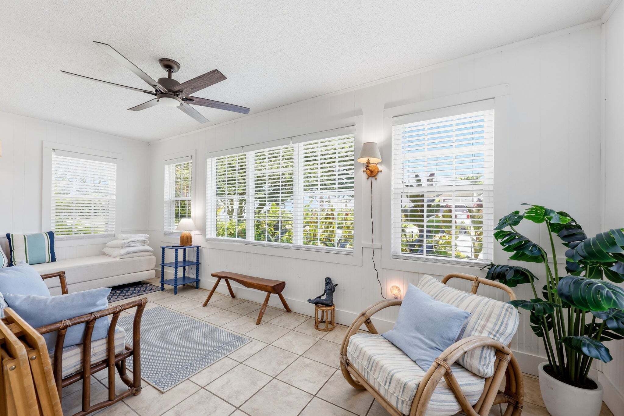A bright room with a ceiling fan, large windows with blinds, wicker furniture, and a potted plant.