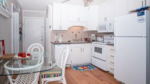 A compact kitchen with white cabinets, a microwave, a refrigerator, and a dining area with a glass table and wicker chairs. - Beautifully Remodeled, Oceanfront, 3rd-Floor Condo with Balcony & Shared Pools (Atlantic Beach)