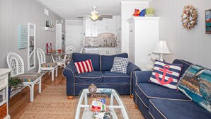 A living room with a dark blue sofa, white wicker chairs, a glass coffee table, and a ceiling fan. - Beautifully Remodeled, Oceanfront, 3rd-Floor Condo with Balcony & Shared Pools (Atlantic Beach)