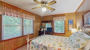 A bedroom with wooden paneling, a bed with a colorful fish-patterned bedspread, a ceiling fan, and large windows with blinds. - Perfect 3BR Getaway with Central A/C, Full Kitchen and Relaxing Balcony (Emerald Isle)