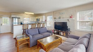 Ein Wohnzimmer mit grauem Sofa, einem Glastisch und einem Fernseher. Eine Küchenzeile mit weißen Schränken und einer Mikrowelle ist ebenfalls vorhanden.