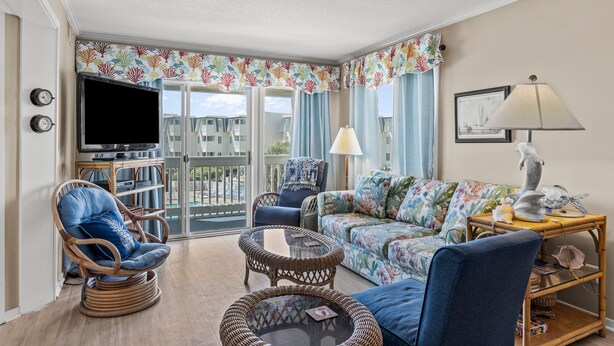 Un salon spacieux avec un balcon, des meubles en osier, un canapé recouvert d’un tissu à motifs floraux et un téléviseur.