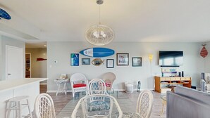 A modern living room with a dining area, white wicker furniture, a large flat-screen TV, and various wall decorations including surfboards and framed pictures.