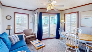 A cozy living room with a blue sofa, a wooden coffee table, a brown armchair, and a dining table with white chairs. - First-Floor Condo with Balcony, WiFi & Shared Pools, Hot Tub, Tennis, Putt-Putt (Atlantic Beach)