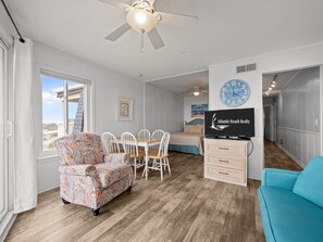 A spacious room with a wooden floor, a ceiling fan, a dining table with chairs, a bed, and a television displaying 'Atlantic Beach Realty'.