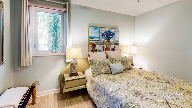 A bedroom with a bed, two bedside tables, a ceiling fan, a window with curtains, and a painting on the wall.