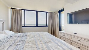 A bedroom with a large bed, a dresser, a flat-screen TV, and a view of the sea through a large window. - Stunning Ocean Front Condo with Shared Amenities | Pool, Tennis, and Gym (Atlantic Beach)