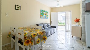 Un coin repas avec une table recouverte d’une nappe à motifs de tournesols, un canapé gris et un meuble blanc orné d’un vase de fleurs.