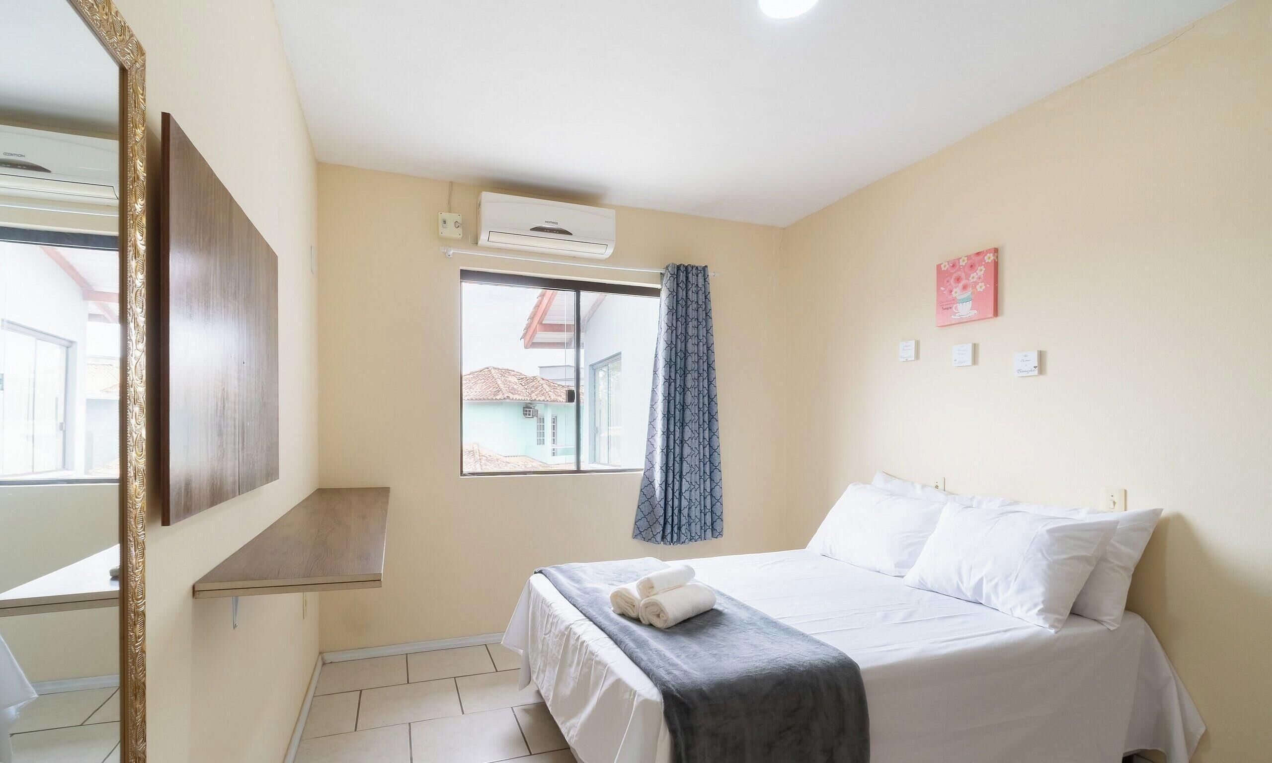 Une chambre d’hôtel avec un lit, une fenêtre avec des rideaux, un climatiseur fixé au mur et une petite étagère.