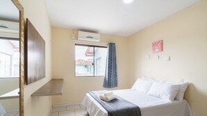 Une chambre d’hôtel avec un lit, une fenêtre avec des rideaux, un climatiseur fixé au mur et une petite étagère.