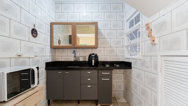 Une cuisine compacte avec des murs recouverts de carreaux blancs, un plan de travail noir, un four encastré et un meuble en bois surmonté d’un miroir.