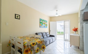 A dining area with a table covered in a sunflower tablecloth, a grey sofa, and a white cabinet with a vase of flowers. - Casa c/ Churrasqueira em Ingleses RIS0006 (Florianópolis)