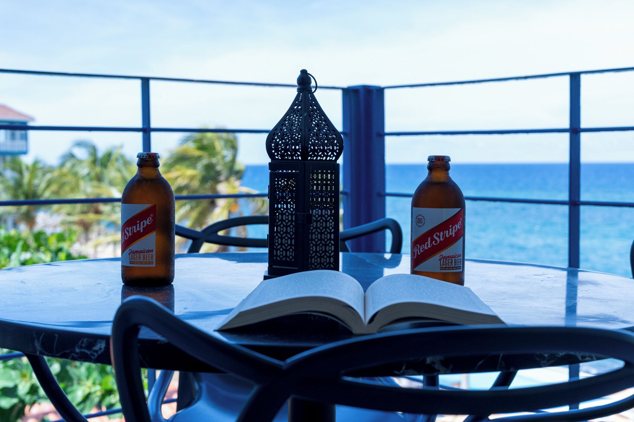 Een tafel met twee Red Stripe biertjes, een boek dat open ligt, en een decoratieve lantaarn op een balkon met uitzicht op de oceaan.