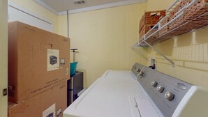 Un área de lavandería con cajas de cartón apiladas, una lavadora y un estante metálico con cestas.