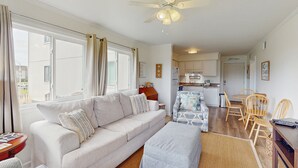 A spacious living area with a sofa, ottoman, and dining set. There is a ceiling fan, a kitchen in the background, and a view through large windows. - Remodeled condo with pool, WiFi, W/D & AC - next to state park (Atlantic Beach)