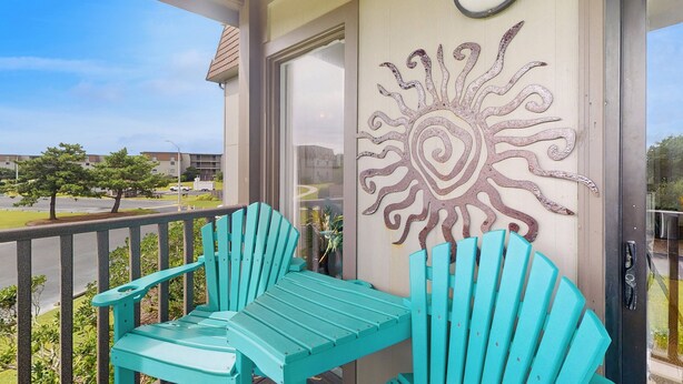 A balcony with turquoise Adirondack chairs, a decorative sun wall, and a view of a parking lot and buildings. - Remodeled condo with pool, WiFi, W/D & AC - next to state park (Atlantic Beach)
