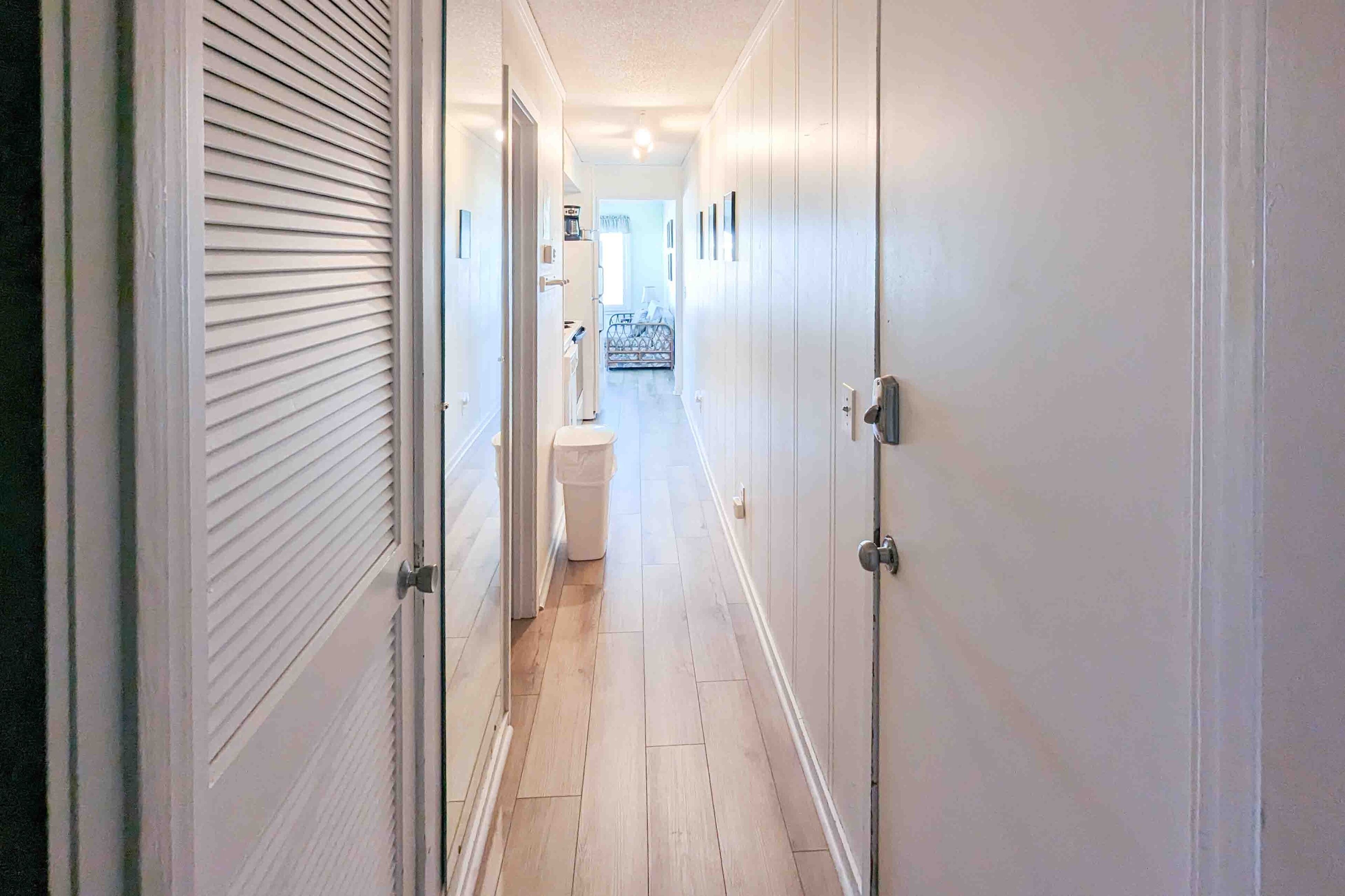 A narrow hallway with white walls and wooden flooring, leading to a bathroom with a toilet and a shower area.